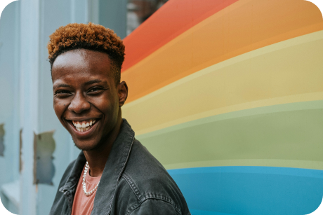 Persona LGBT sorridente in posa davanti a un murale arcobaleno.